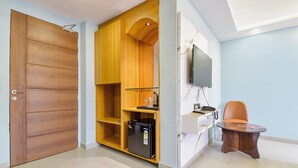 A room with a wooden wardrobe, a small kitchen area with a refrigerator, a television mounted on the wall, and a single orange chair. - Fabhotel Pink Palace Raja Park (Jaipur)
