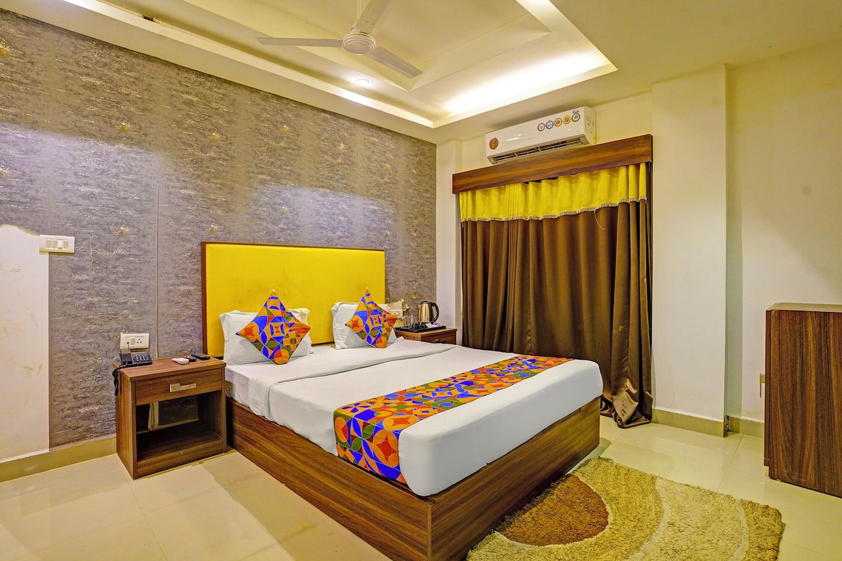 A hotel room with a wooden headboard, a bed with colorful bedding, a nightstand with a phone, and an air conditioning unit on the wall.