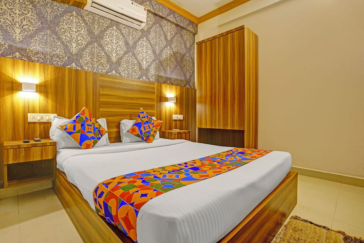 A hotel room with a large bed, wooden headboard, two bedside tables, a nightstand, a wardrobe, and an air conditioning unit.