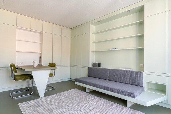 A modern, compact room with a grey sofa, a small table, and a chair. There are built-in shelves on the walls.