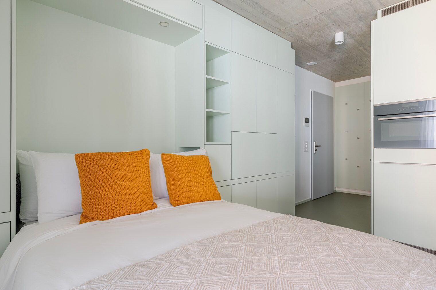 A bedroom with a bed, two pillows, a wardrobe, and a kitchenette.