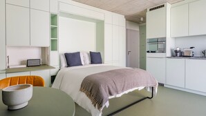 A modern, compact hotel room with a bed, a small table with a bowl, a chair, a kitchenette, and a wardrobe.