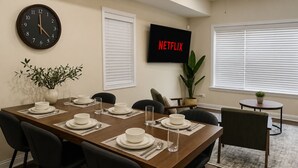 Comedor con mesa de madera dispuesta para cuatro, sillas, un reloj, un televisor mostrando 'NETFLIX' y una planta en maceta.