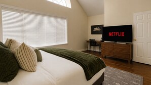 Un dormitorio con cama, escritorio, silla, televisor mostrando Netflix, puerta y ventana con persianas.