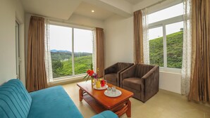 Una habitación amplia con un ventanal grande, un sofá azul, un sillón marrón y una mesa de centro de madera con un jarrón de flores y una bandeja de vasos.