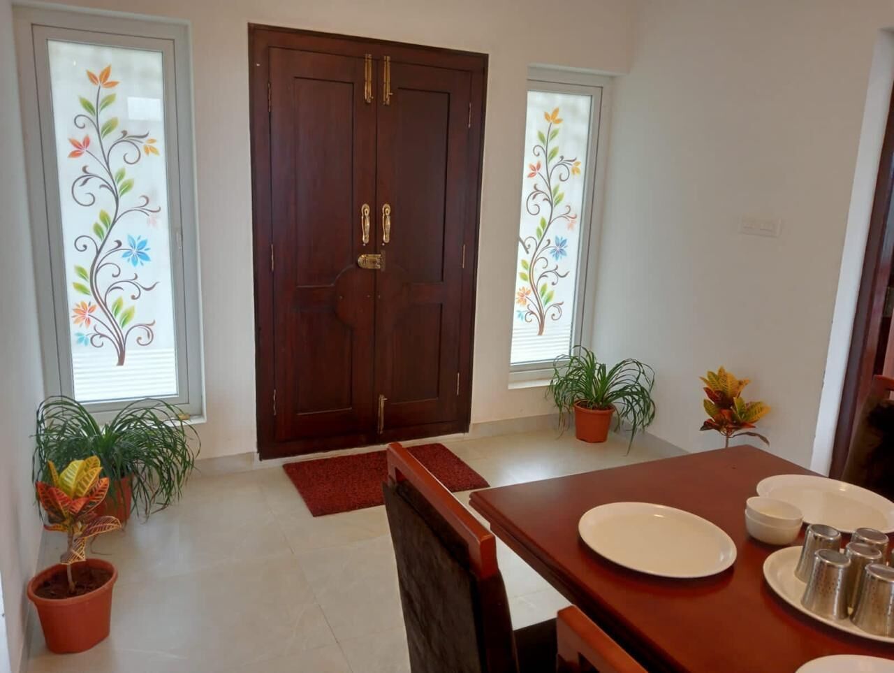 Un coin repas avec une table en bois dressée pour quatre personnes, deux plantes en pot et une grande porte en bois dotée de panneaux de verre.