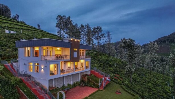 Una casa moderna de dos pisos con una escalera cubierta de alfombra roja, rodeada de abundante vegetación y colinas.