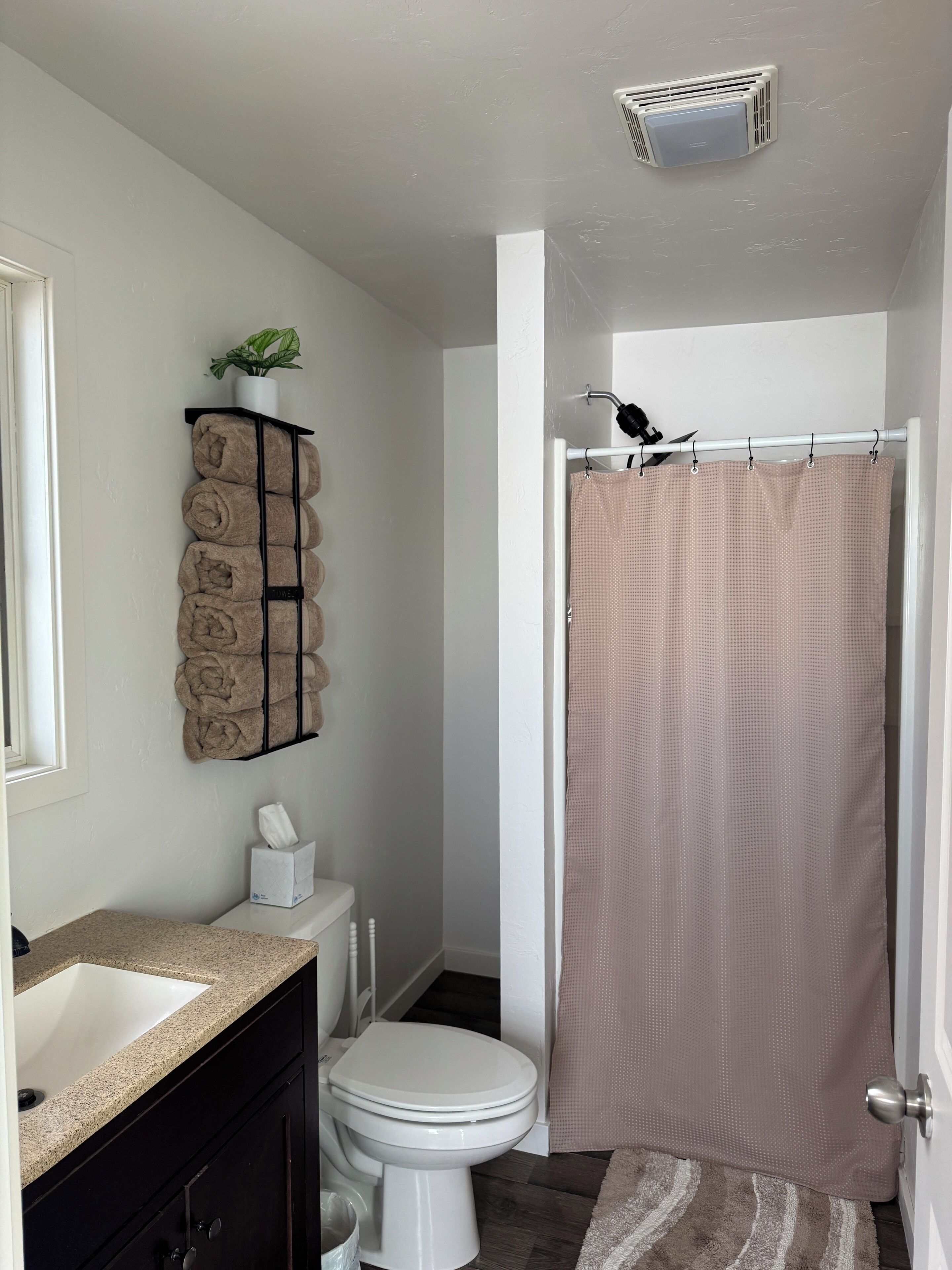 A bathroom with a white toilet, a sink, a towel rack with rolled towels, a shower curtain, and a ceiling vent.