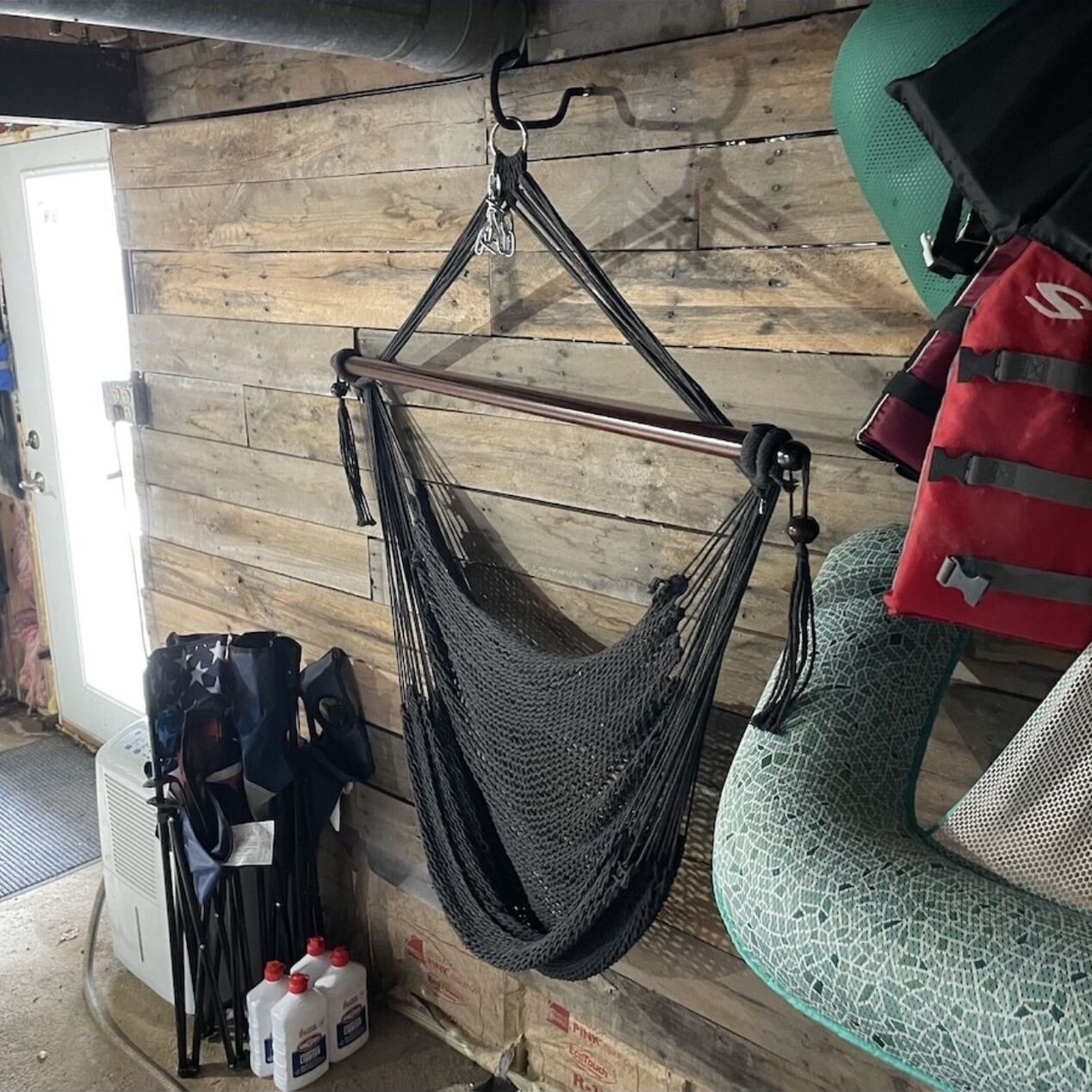 A hammock suspended from a ceiling in a room with wooden walls, a green patterned chair, and a red bag with a white logo.