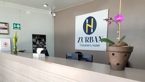 A hotel reception desk with a sign for ZURBAN Travelers Hotel, a potted orchid, and a display of snacks. - ZURBAN TRAVELERS (Paraíso)