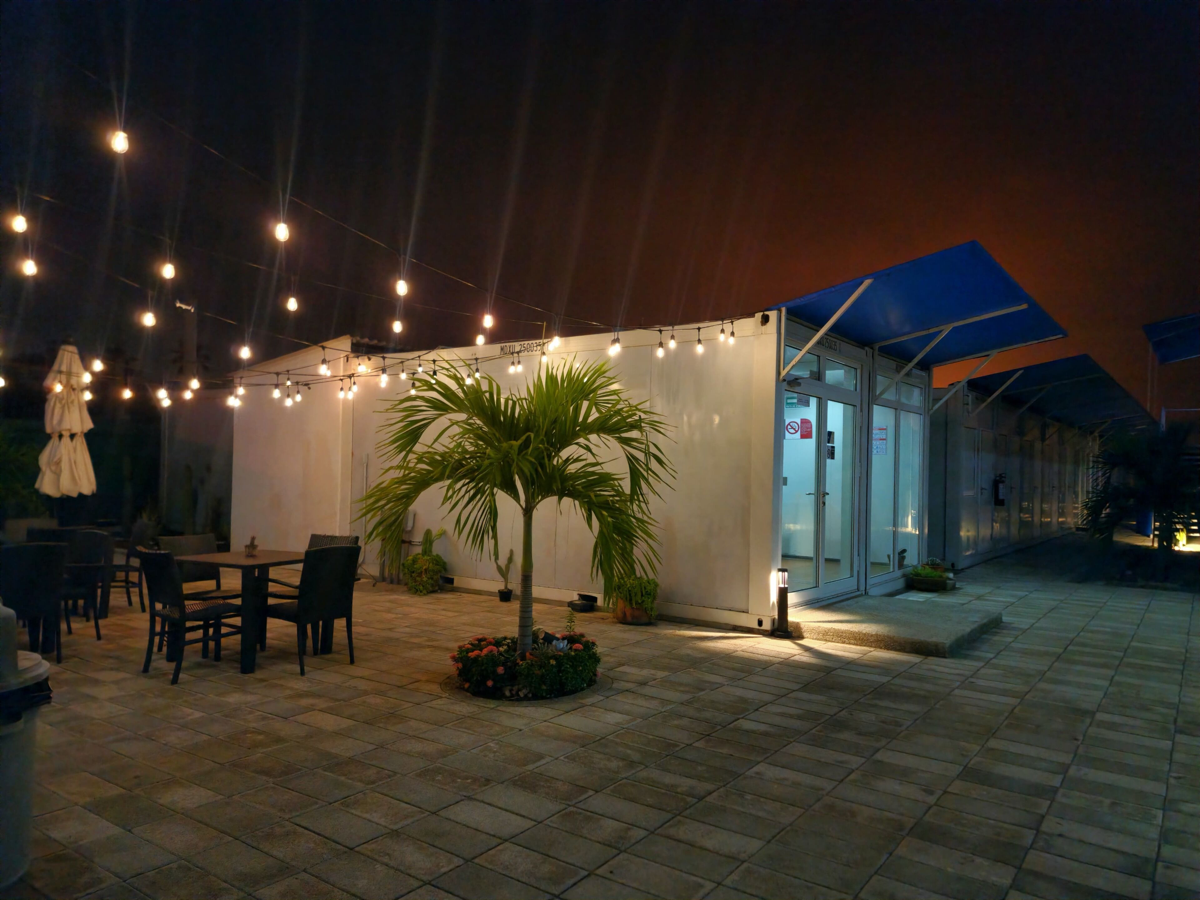 Un patio al aire libre con mesa y sillas, luces colgantes y una palmera.