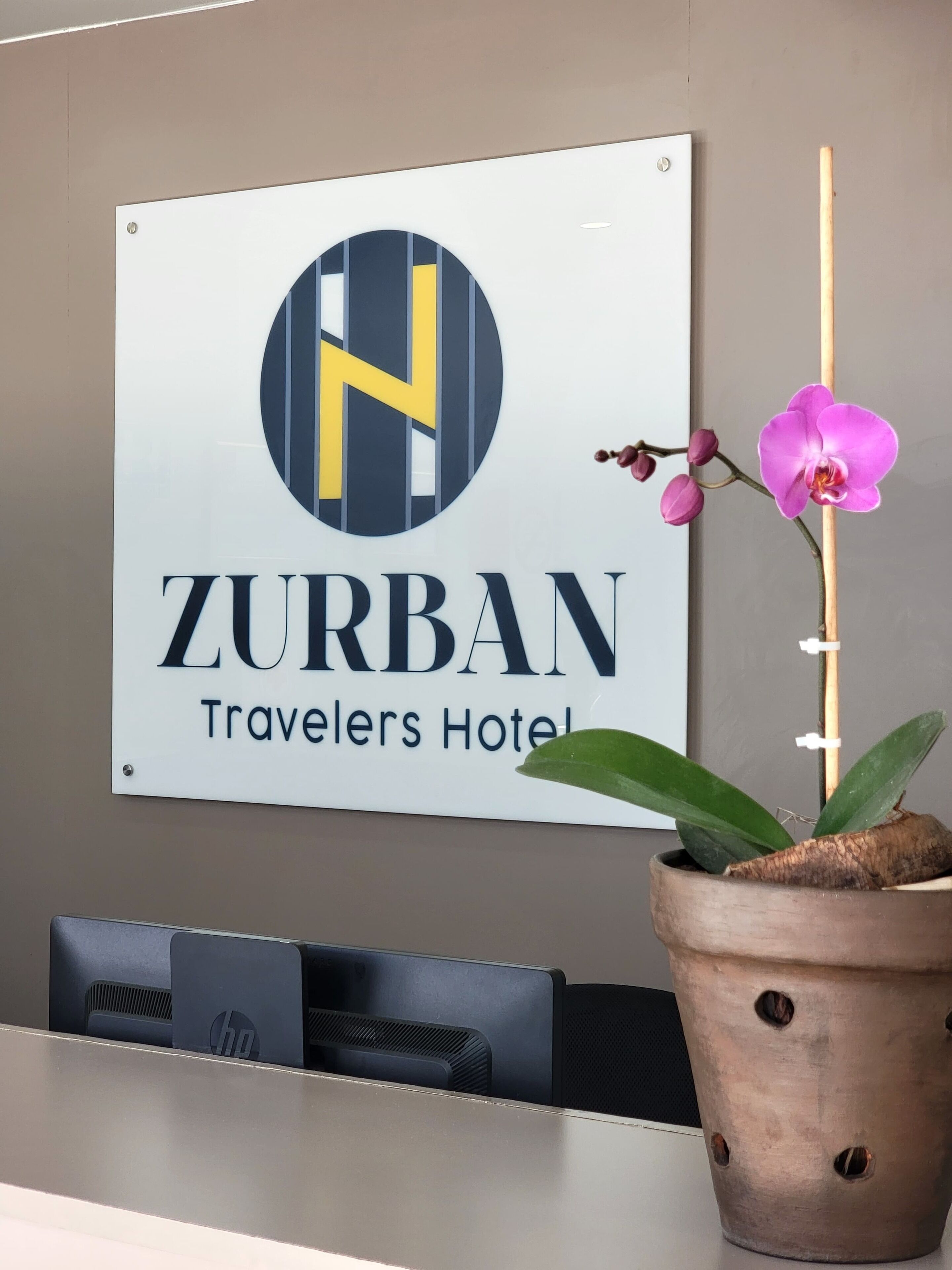 A sign for ZURBAN Travelers Hotel with a logo, a reception desk, and a potted orchid.