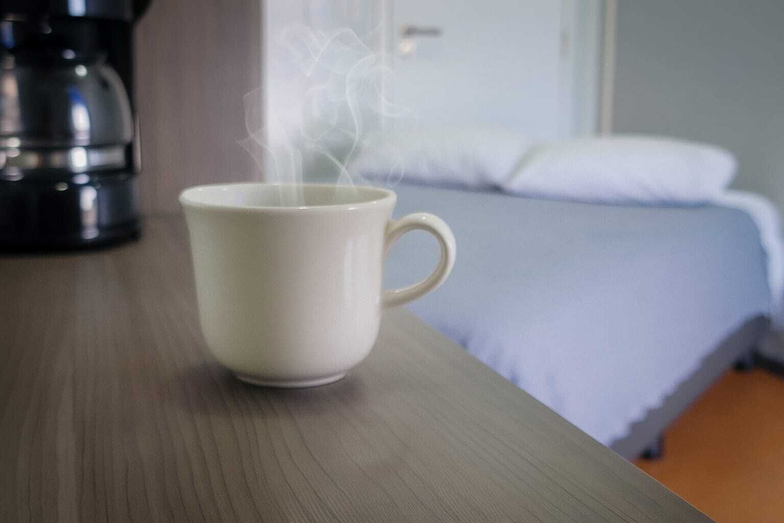 A white mug on a wooden table with steam rising from it, a bed with a white pillow and blue bedding in the background, and a coffee maker to the left.