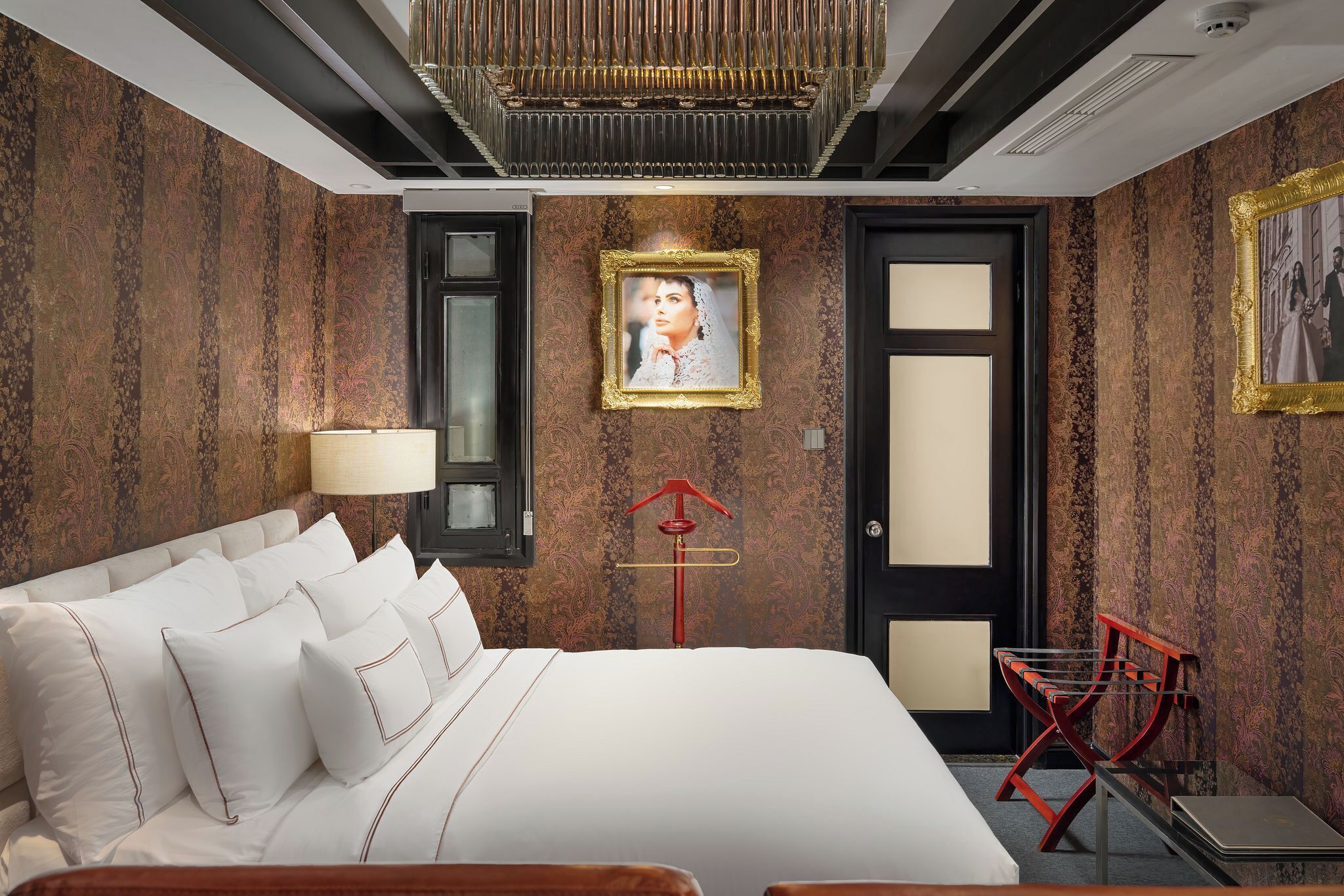 Une chambre d’hôtel avec un grand lit, une lampe de sol rouge, un tableau encadré au mur et une porte à cadre noir.