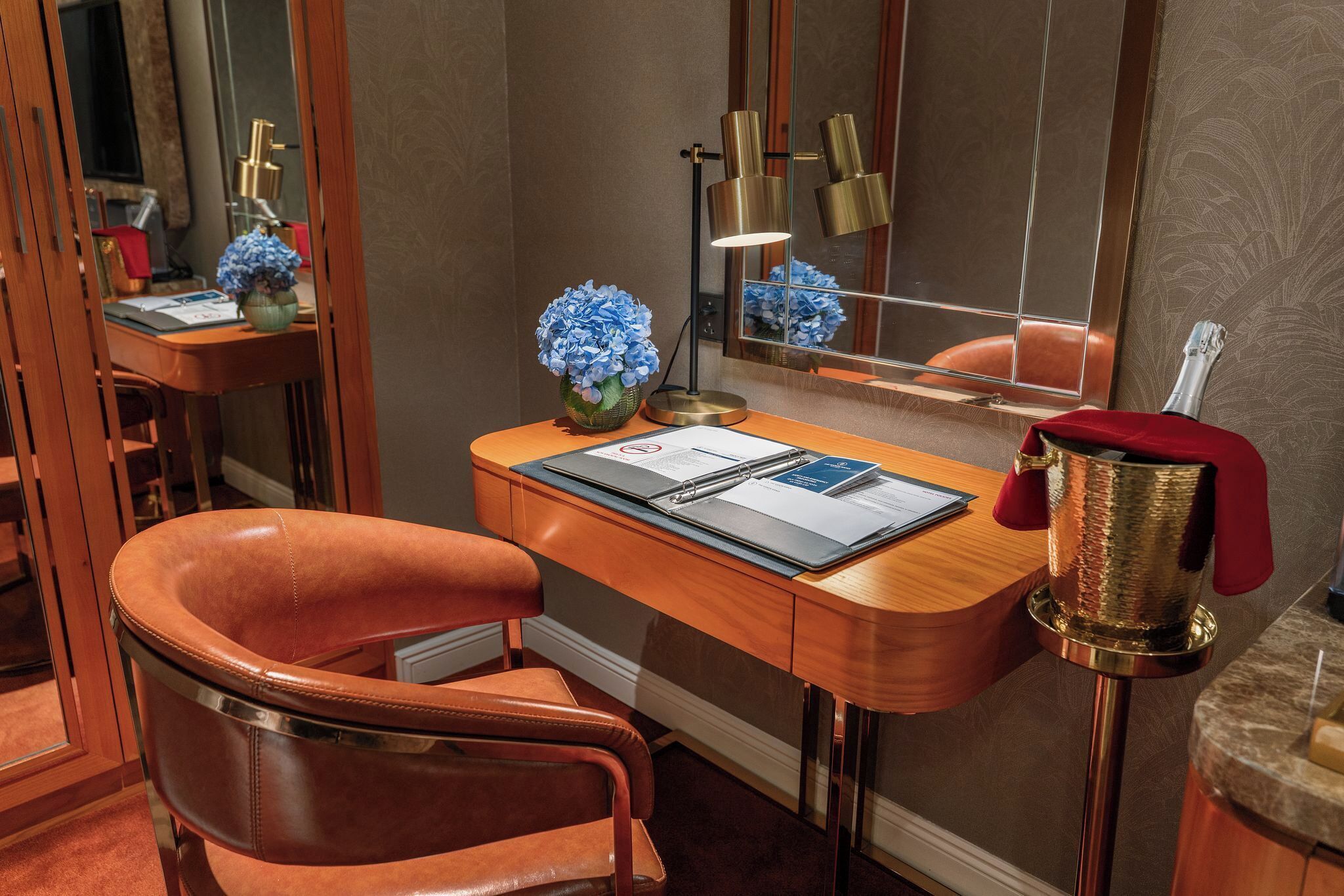 A hotel room with a wooden desk, a leather chair, a mirror, a vase with blue flowers, a lamp, and a bottle of champagne.