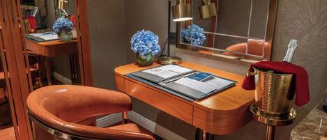 A hotel room with a wooden desk, a leather chair, a mirror, a vase with blue flowers, a lamp, and a bottle of champagne.