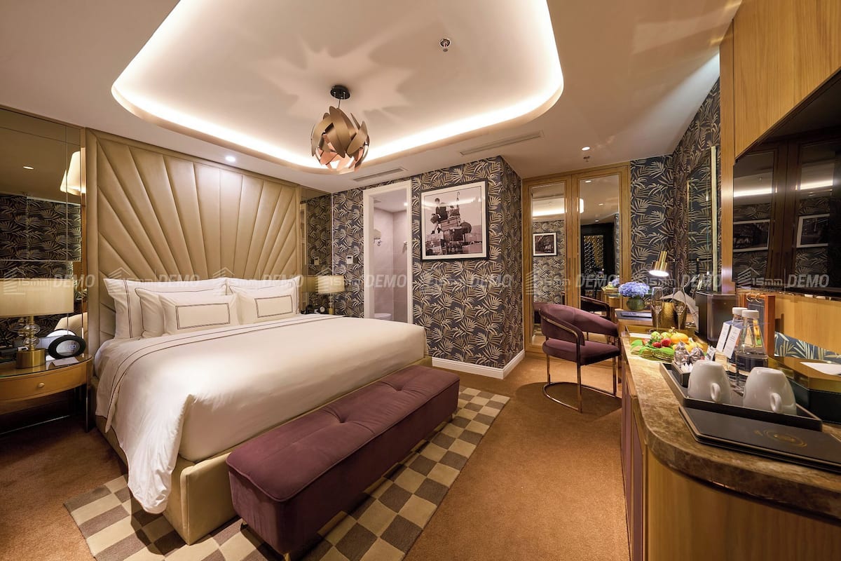 A modern hotel room with a large bed, a bench, a desk with a laptop, and a minibar.