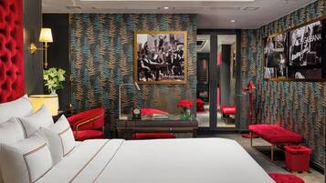 Camera d'albergo moderna con un letto grande, poltrone rosse rivestite in tessuto, una scrivania con una lampada e un quadro appeso al muro.