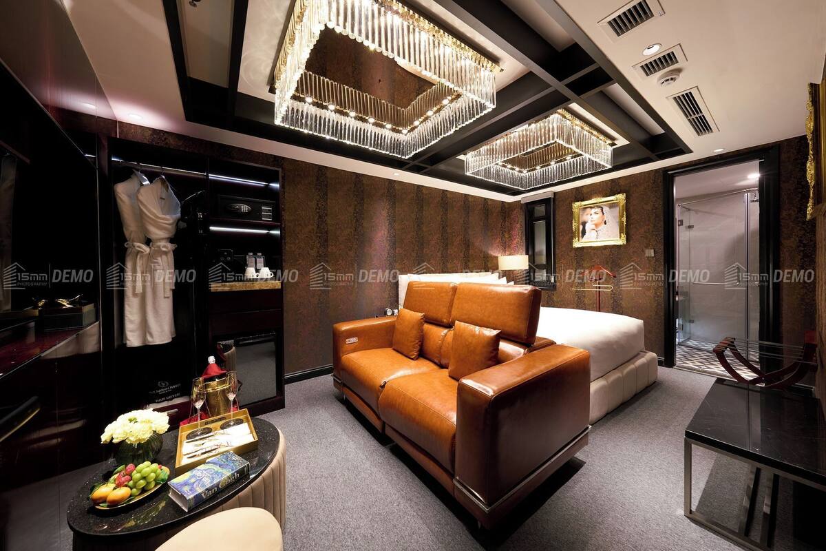 A modern hotel room with a leather sofa, a bed, a vanity with a mirror, and a chandelier.