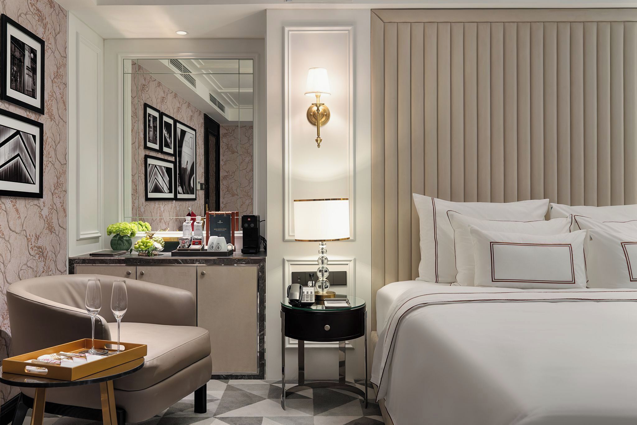 A hotel room with a bed, a desk with a lamp, a chair, and a tray with glasses.