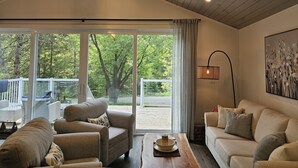 Un salón espacioso con un amplio ventanal corredera que da a una terraza exterior, techo de madera y una lámpara de pie moderna.