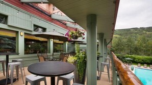 An outdoor patio with round tables and chairs, a hanging flower pot, and a view of a swimming pool and forested hills. - Convenient 2BR Condo Near Lionshead Village With Resort Amenities (Vail)