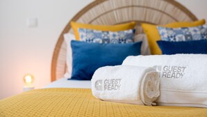 A neatly made bed with a yellow bedspread and a rolled white towel with "GUEST READY" embroidered on it.