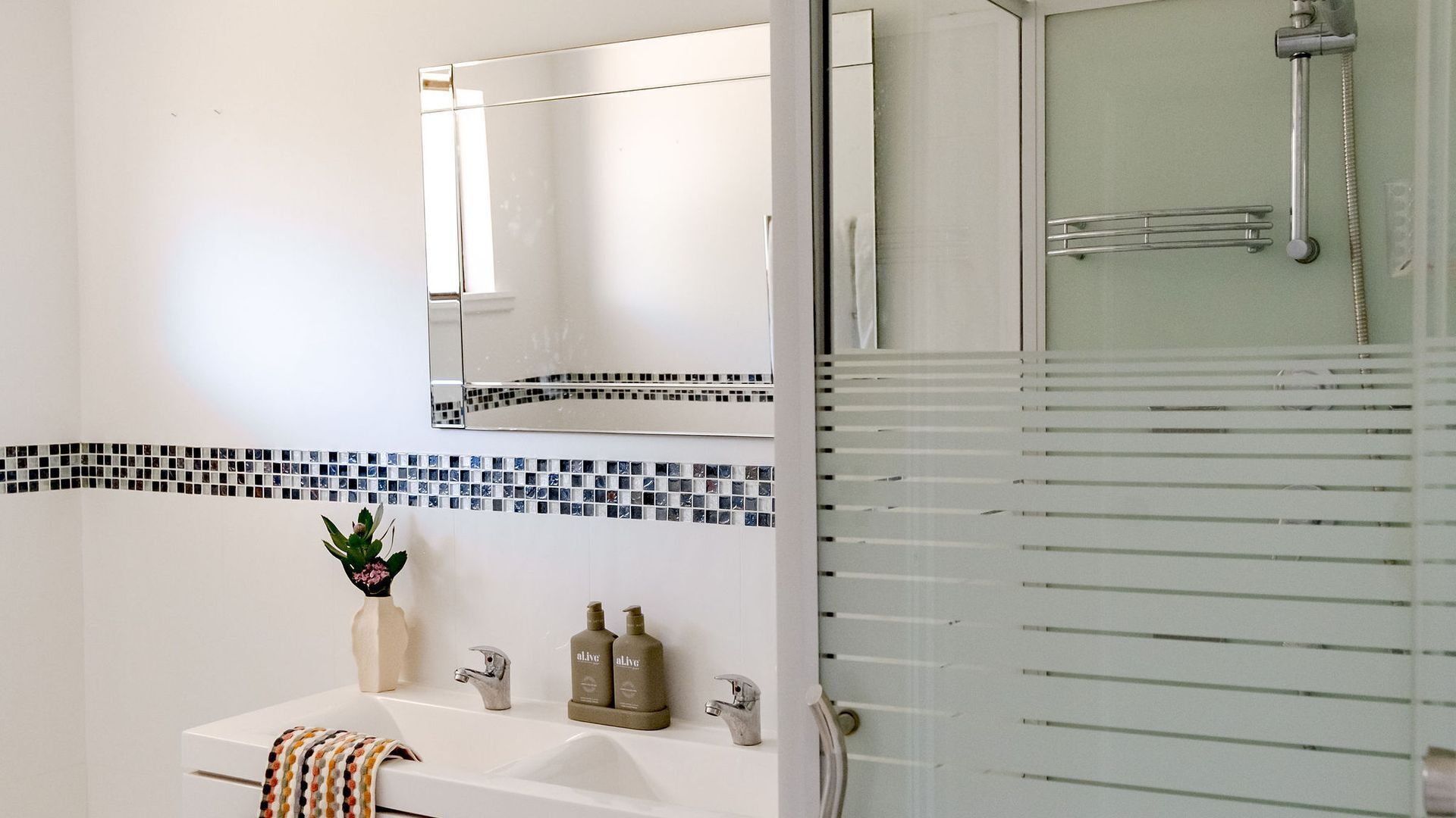 A bathroom with a white sink, two bottles of lotion, a vase with a plant, and a shower with a glass door.