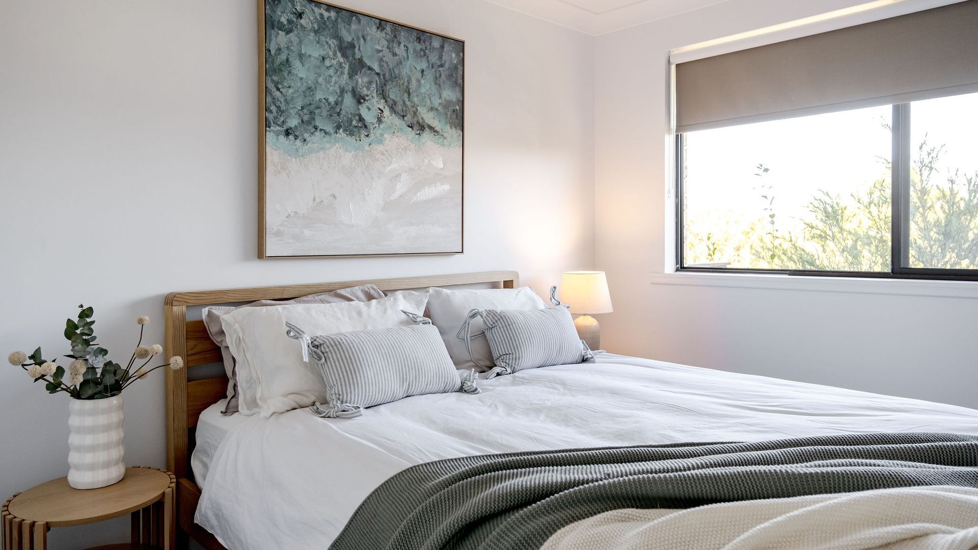 A neatly made bed with white linens and striped pillows, a bedside table with a vase of flowers, and a large abstract painting above the bed.