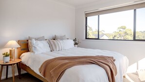 A bedroom with a bed, a bedside table with a lamp, a window with a view of trees, and a small plant on the table.