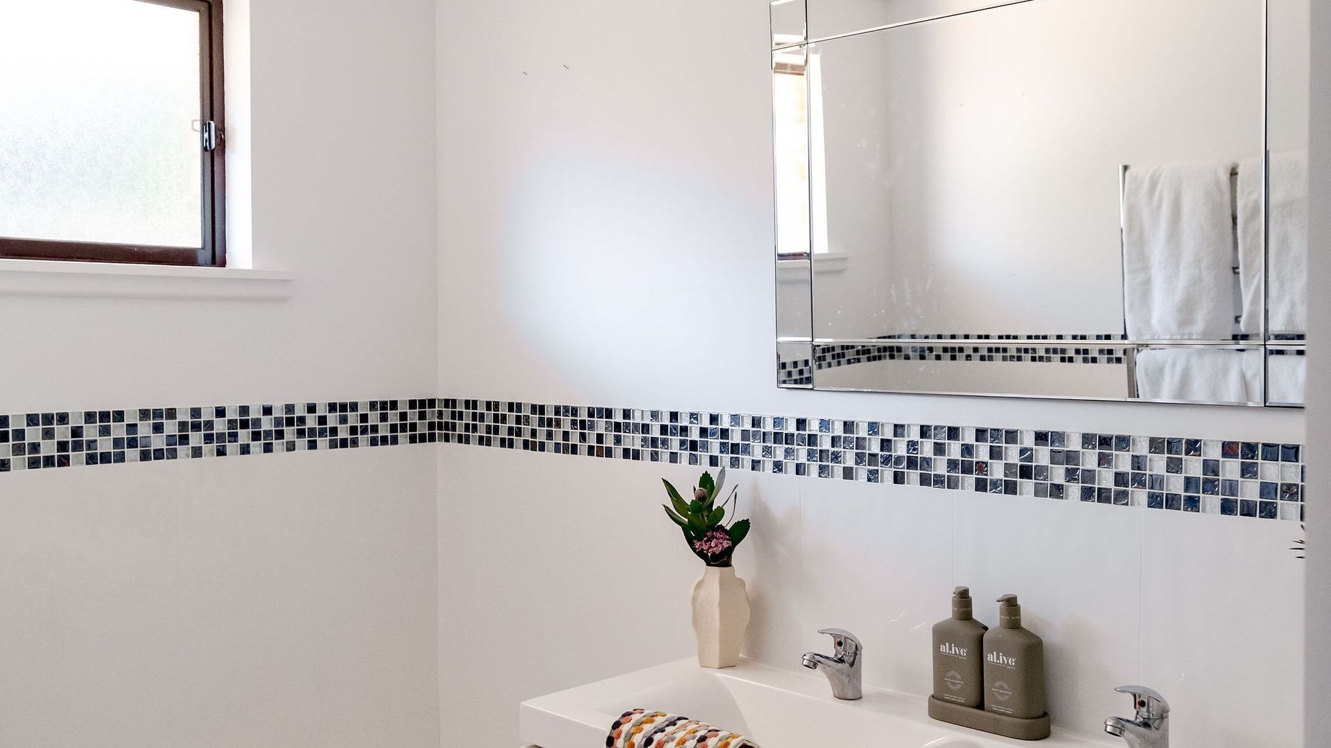 Un baño con lavamanos blanco, dos llaves, un espejo sobre el lavamanos y una ventana a la izquierda.