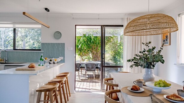 A modern kitchen with a large island, wooden stools, a dining table with a vase of flowers, and a view of the outdoors through a glass door.