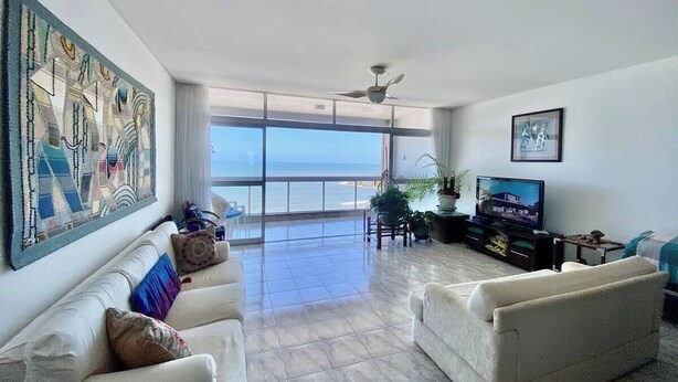 A spacious living room with a large window overlooking the sea, a ceiling fan, and a television on a stand. - A Beachfront Oasis, 220m (Guarujá)