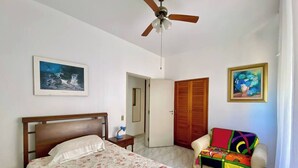 A bedroom with a bed, a ceiling fan, a door, a chair, and two paintings on the wall. - A Beachfront Oasis, 220m (Guarujá)
