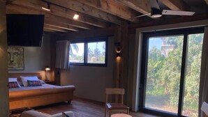 Habitación con sofá, dos sillones, una mesita, televisor montado en la pared y ventanas amplias con vistas a zonas verdes.