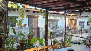 Un espace extérieur aménagé, abrité et doté de tables et de chaises, entouré de plantes en pot et d’un mur de pierre.