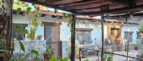 Un área de estar al aire libre cubierta, con mesas y sillas, rodeada de plantas en macetas y un muro de piedra.