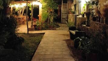 Een geplaveid pad leidt naar een houten terras met buitenmeubilair, omgeven door potplanten en een stenen muur.