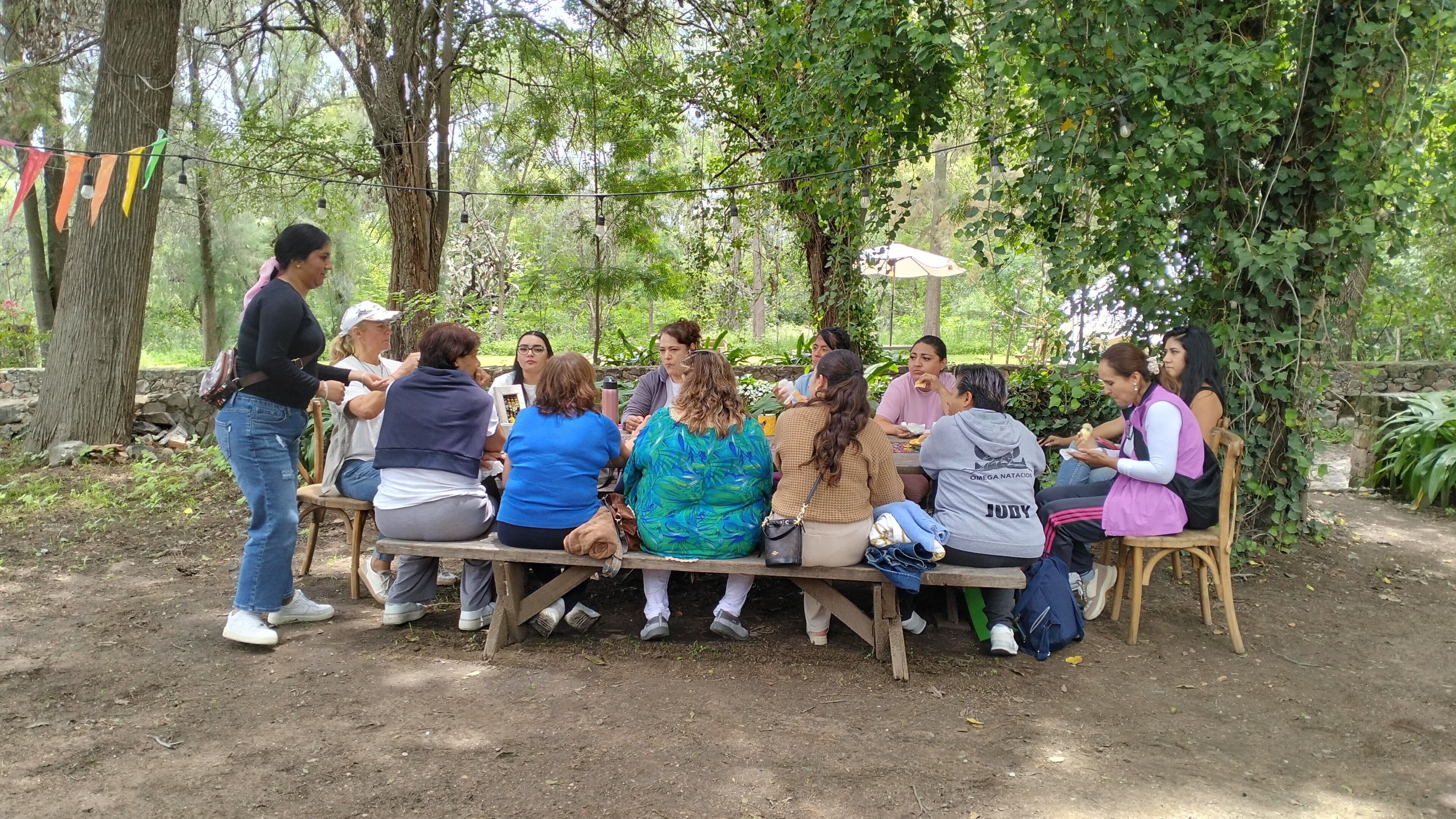 A group of people seated on a wooden bench outdoors, surrounded by trees and greenery.