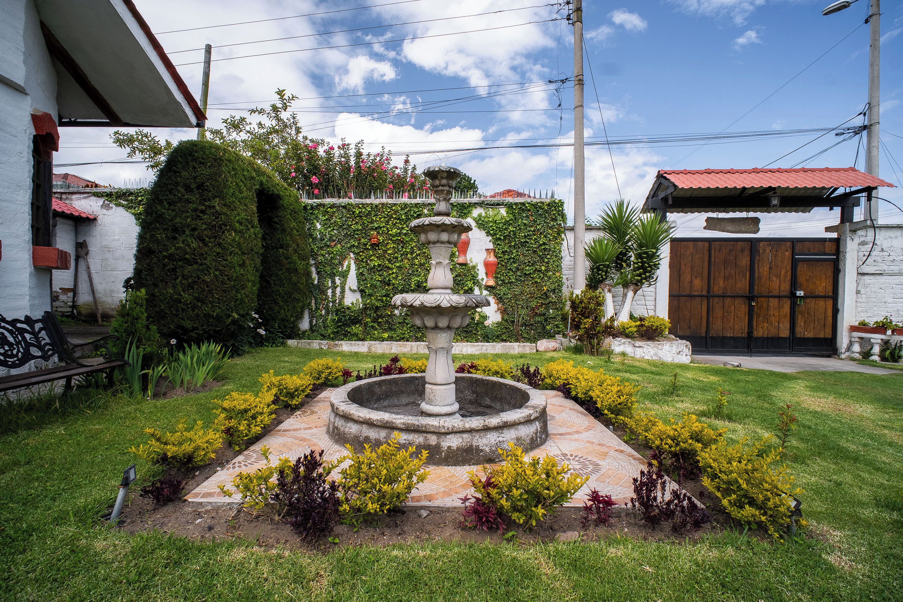 A garden with a central fountain, a white building with a red roof, and a wooden door.