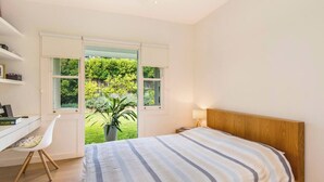 Ein Schlafzimmer mit Bett, Schreibtisch und Stuhl. Zwei Fenster bieten Blick auf Grünflächen.
