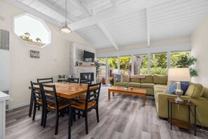 A spacious living area with a wooden dining table, chairs, a green sofa, and a coffee table. A fireplace is built into the wall, and there is a large window providing a view of the outdoors. - 3 Bedroom Home - Walking distance to beach! (Aptos)