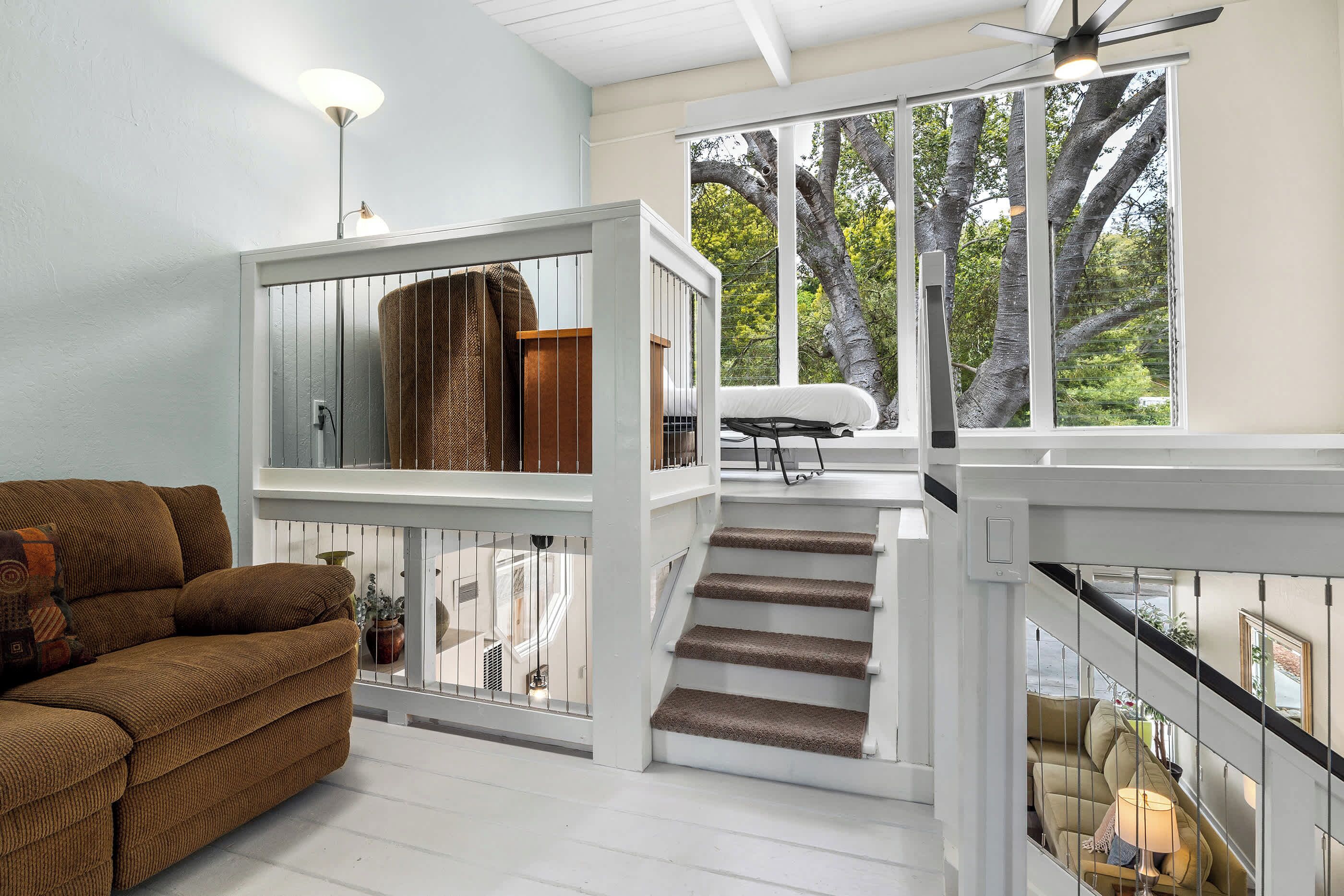 A spacious living area with a staircase, a sofa, and a view of trees through large windows.