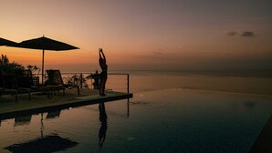 En person laver yoga på en flydende platform ved siden af en pool med liggestole og parasoller.