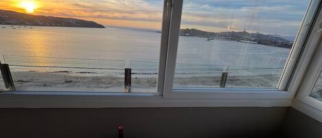 A balcony with a view of the sea at sunset, a table with bottles and glasses, and a bench.