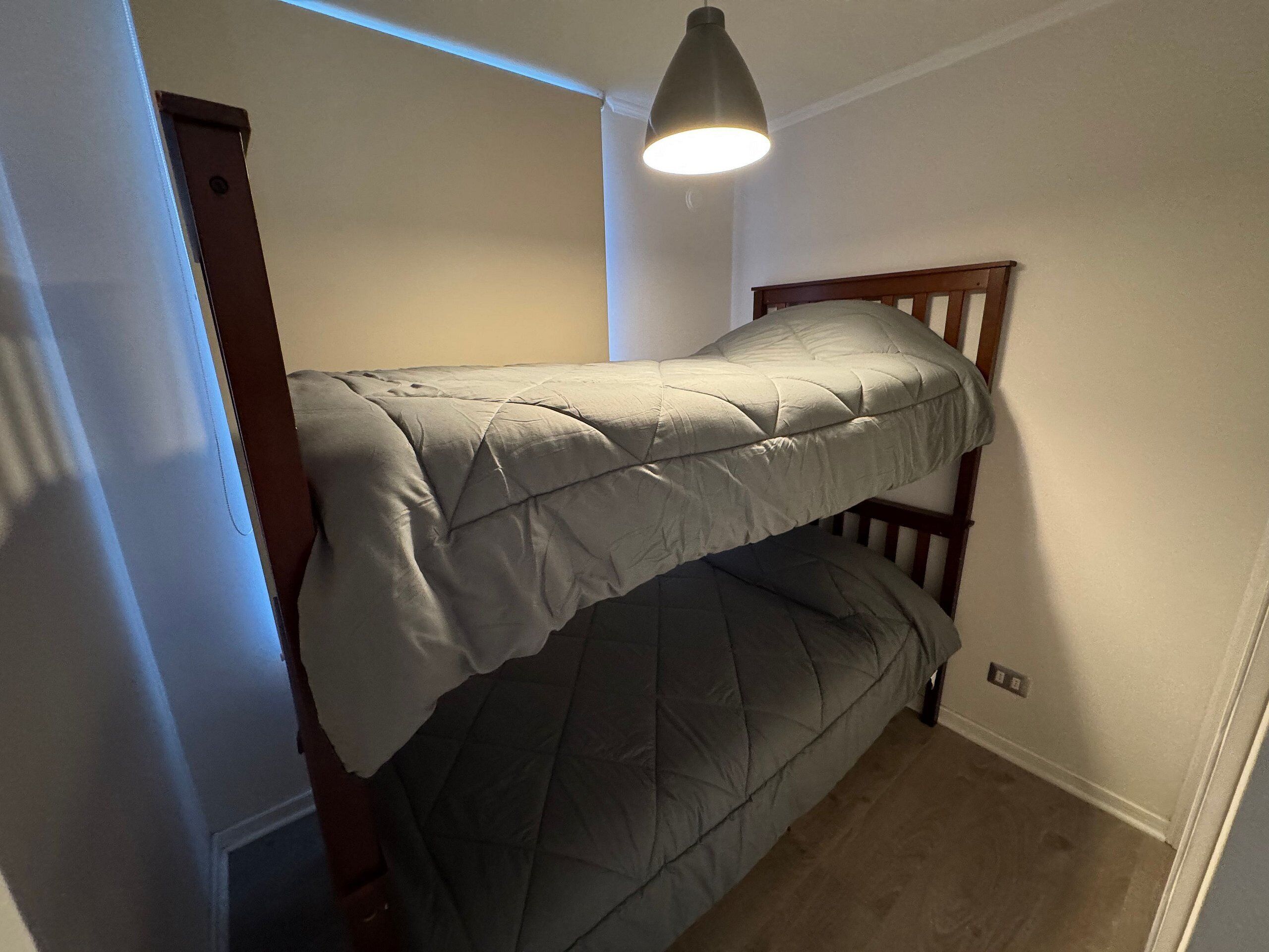 A small, single-bed room with a wooden headboard, a hanging light fixture, and a door to the left.