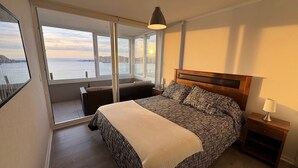 A bedroom with a large bed, a nightstand, a lamp, and a view of the sea through large windows. - Costa Herradura (Coquimbo)