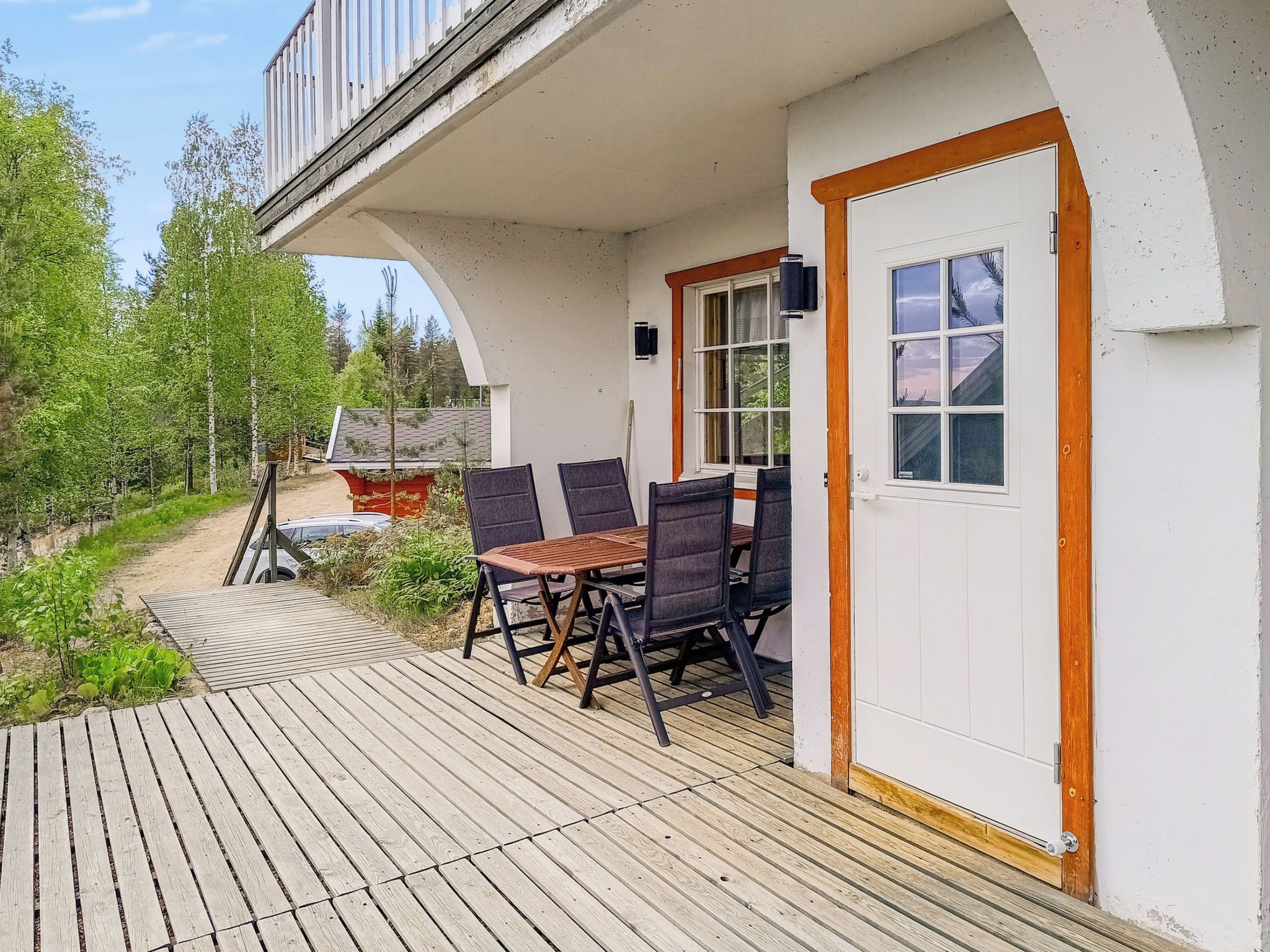 A wooden deck with a table and chairs, a white door, and a view of a forested area.