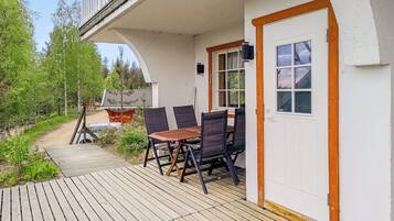 A wooden deck with a table and chairs, a white door, and a view of a forested area.