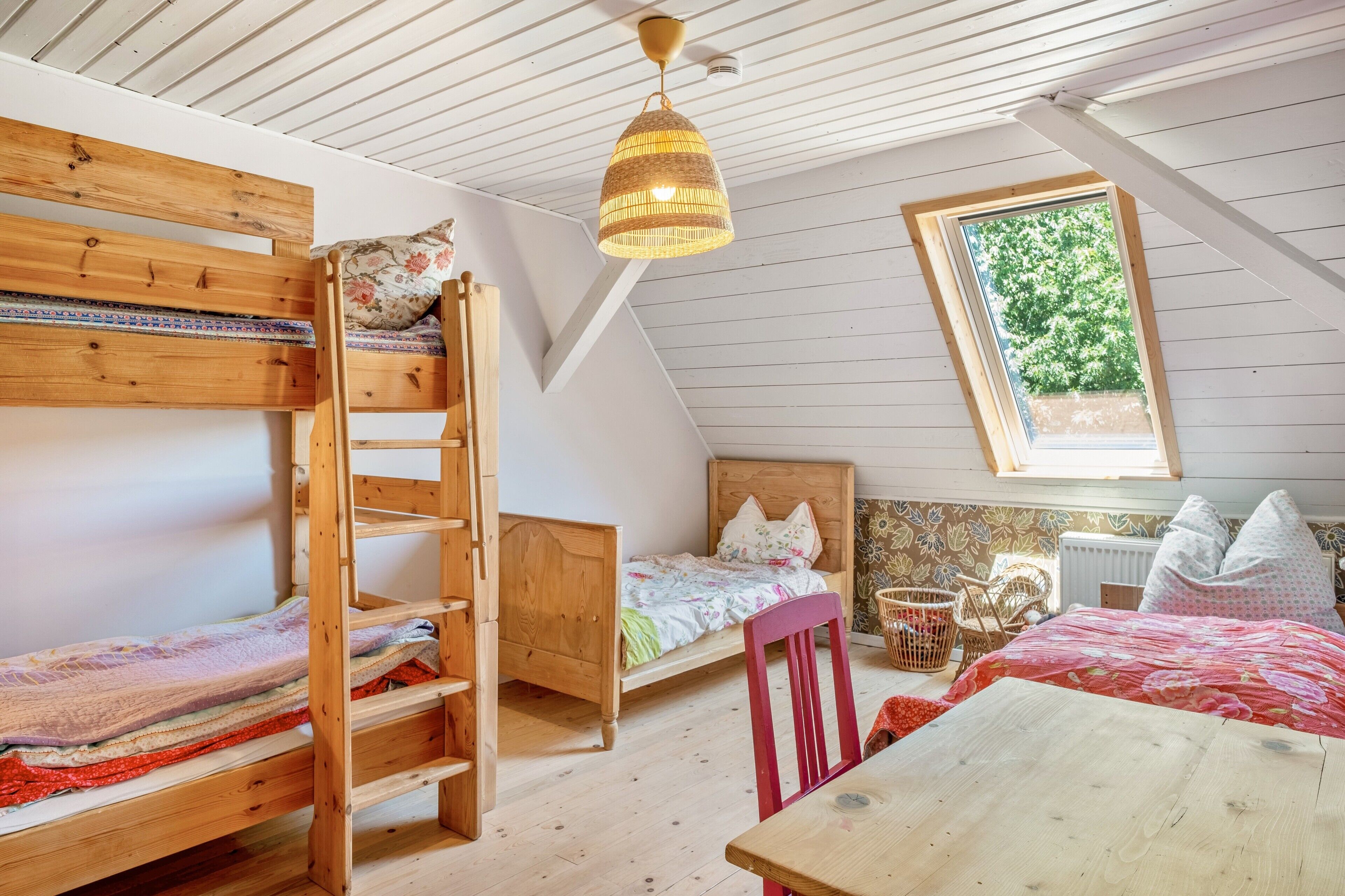Un dormitorio estilo loft con una cama litera, una cama individual, una mesa de madera, una silla de mimbre y un ventanal con vistas a árboles.